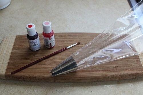 Plastic pasty bag on wood board with two jars of food color and a small paint brush.