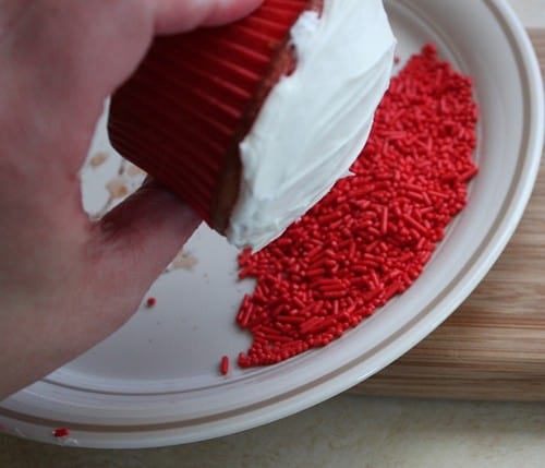 Dipping top of white frosted cupcake in red sprinkles.