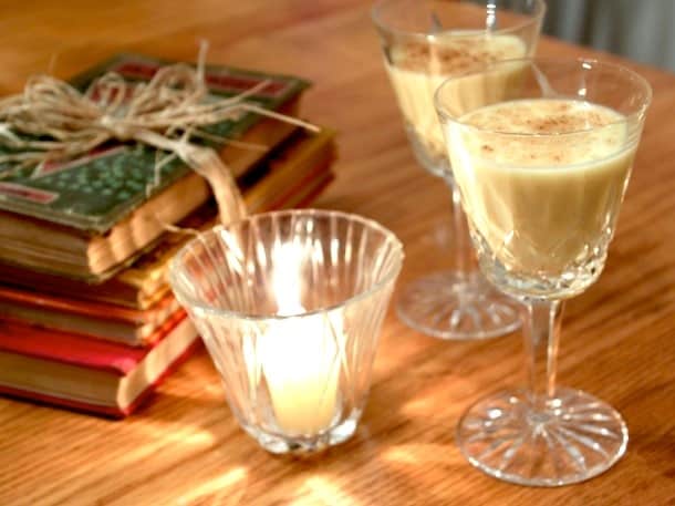 Two glasses of eggnog next to a candle and stack of old books.