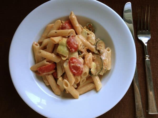 Gluten-free pasta in white bowl with cream sauce, tomatoes, and artichoke hearts.