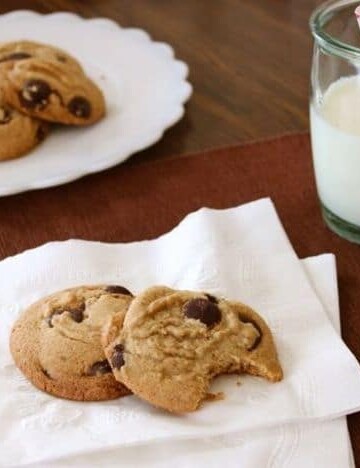 Gluten-free brown butter chocolate chip cookies on a napkin. A glass of milk and a platter of cookies is in the background.