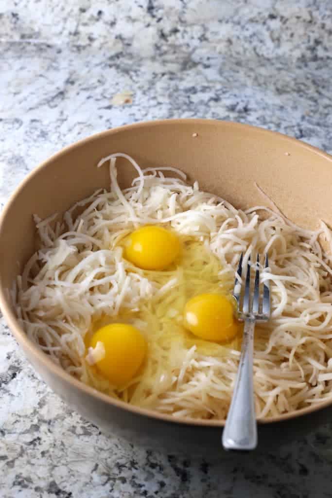 Shredded latkes mixture in brown bowl with three eggs.  