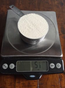 Cup of brown rice flour on a digital scale. Display reads 5 1/8 ounce.