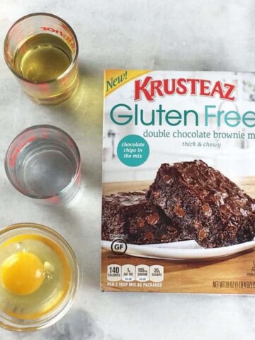 Krusteaz gluten free double chocolate brownie mix box with oil, water, and an egg sitting beside it.
