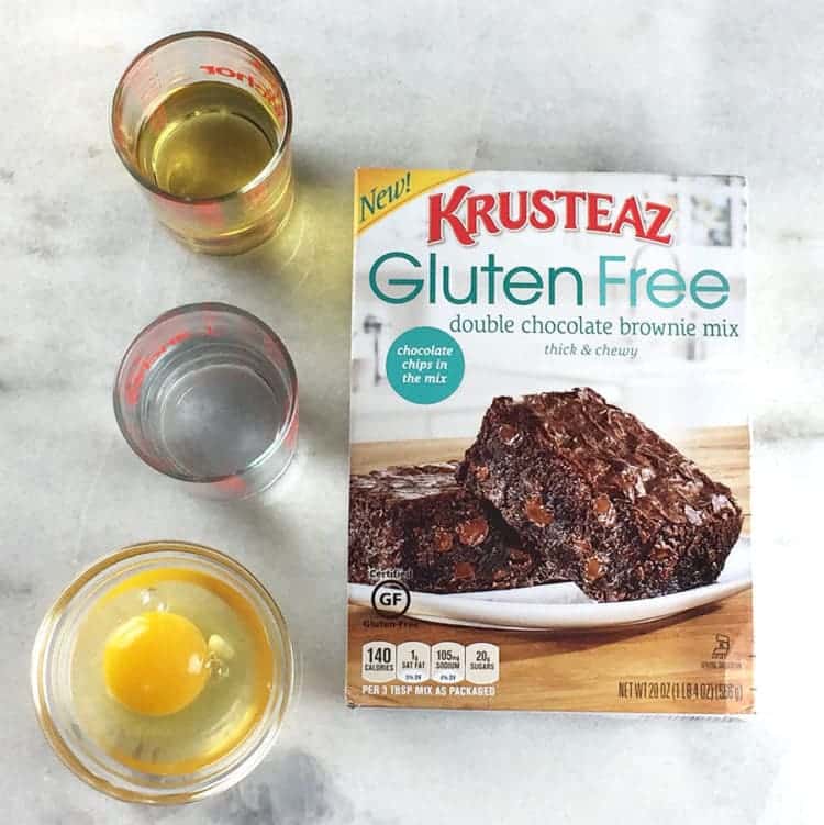 Krusteaz gluten free double chocolate brownie mix box with oil, water, and an egg sitting beside it.