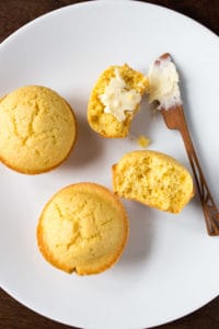 Gluten-free corn muffins on a white plate. One is split and spread with butter.