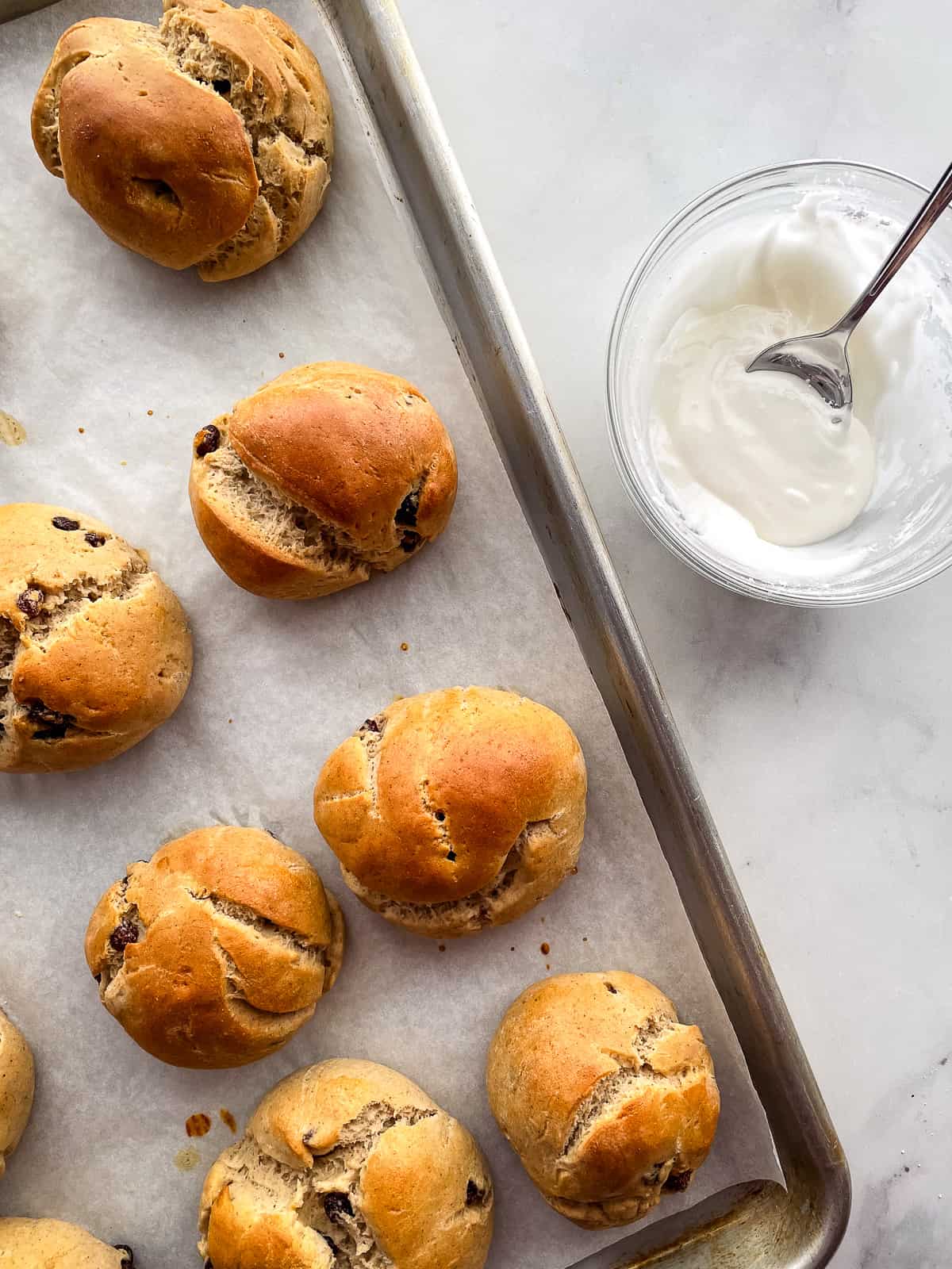 Gluten-free hot cross buns on a baking sheet. A small bowl of icing sits off to the side.