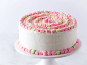 Strawberry cake with white frosting and pink and green marshmallows sits on a cake stand.