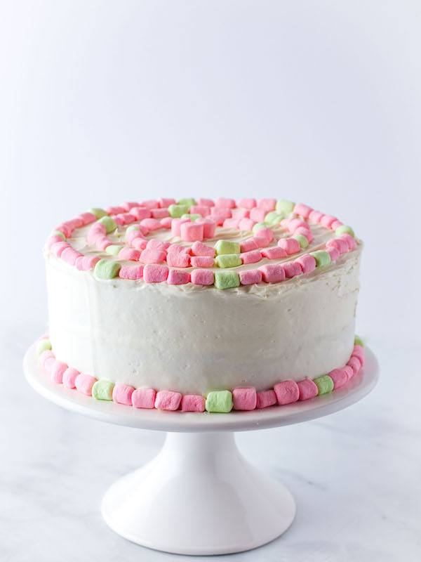 Gluten-Free Strawberry Cake on a white platter. Frosted with cream cheese frosting and decorated with pink and green mini-marshmallows. 