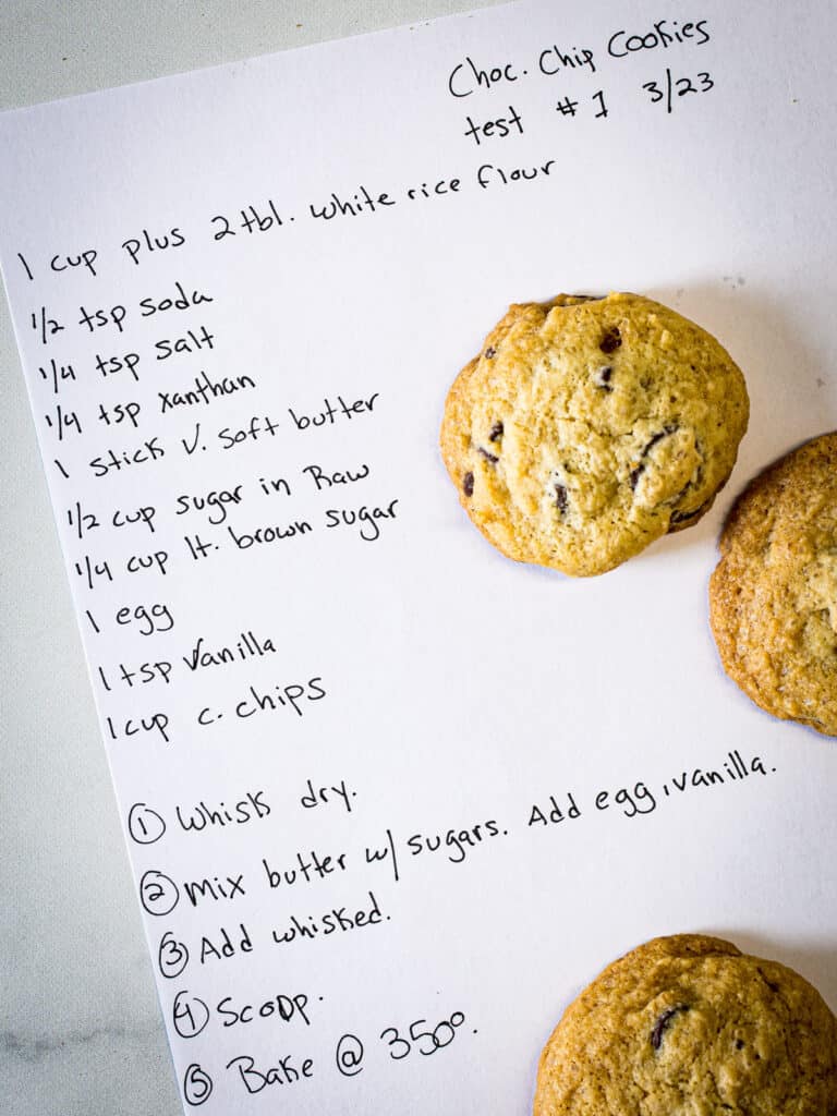 Handwritten gluten-free chocolate chip cookie recipe on paper. Three cookies sit on the paper.