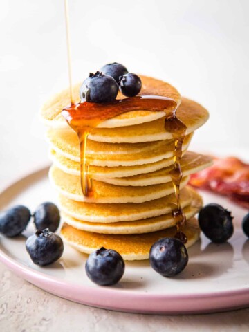 Stack of coconut flour pancakes topped with blueberries and maple syrup on a small pink plate.