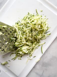 Grated zucchini on a cutting board with a box grater on the left.