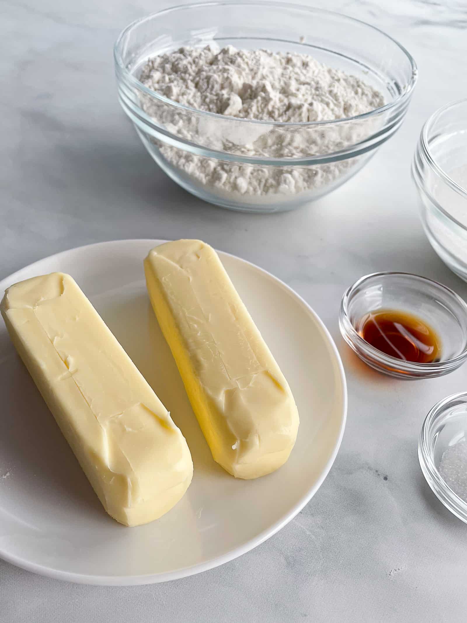 Ingredients for gluten-free shortbread. Two sticks of butter are up front on a plate. Gluten-free flour, sugar, vanilla, and salt are in the background.