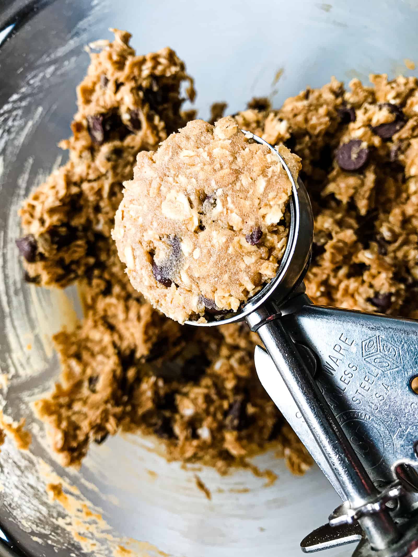 A cookie scoop filled with gluten-free oatmeal cookie dough. A bowl of dough is in the backdround.