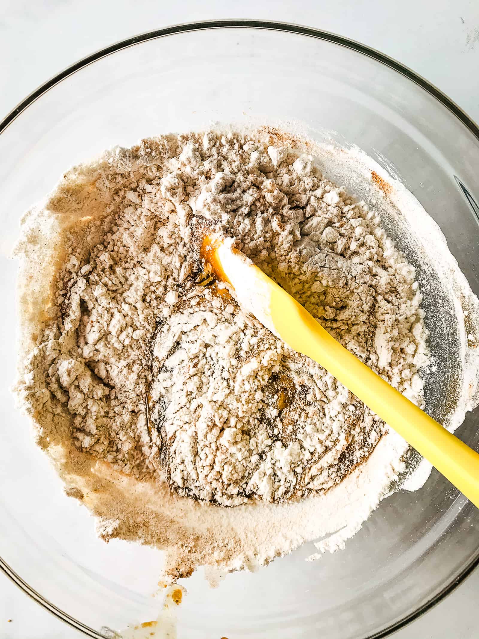 Gluten-free oatmeal cookie dough being mixed in a glass bowl. Gluten-free flour sits ontop of the butter and egg mixture. A yellow spatula is in the bowl.
