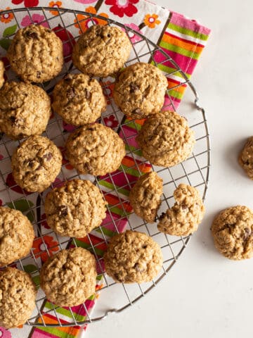 Gluten-free oatmeal cookies cooling on a wire rack. A brightly colored tea-towel sits under the wire rack. Two cookies are on the board off to the right.