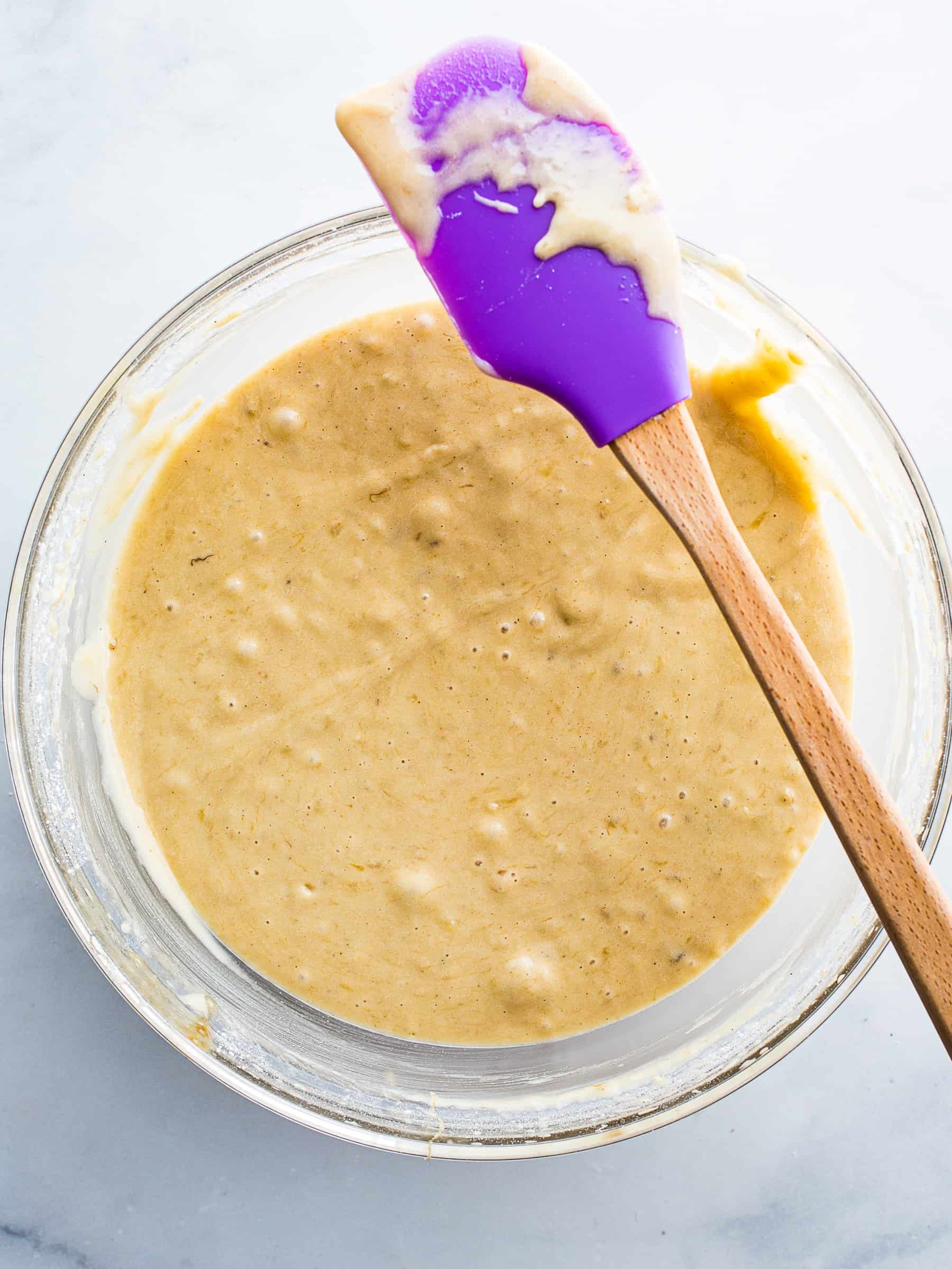 Gluten-free banana muffin batter in a glass bowl. There is a purple spatula sitting on the bowl.