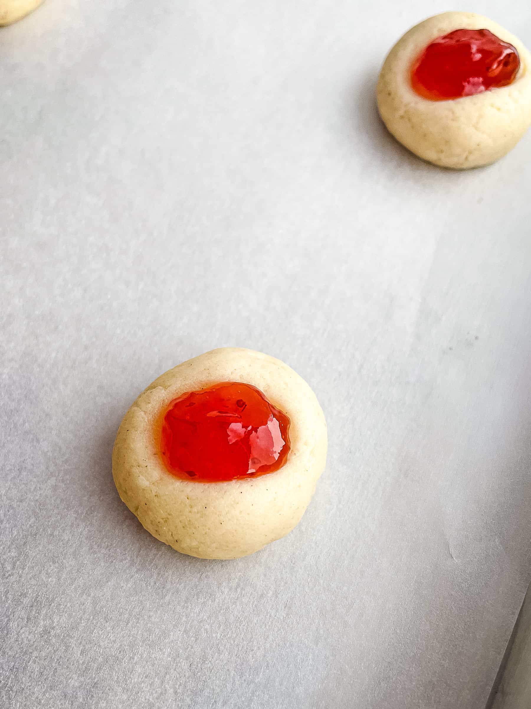 Unbaked gluten-free thumbprint cookies topped with strawberry jam on a baking sheet.