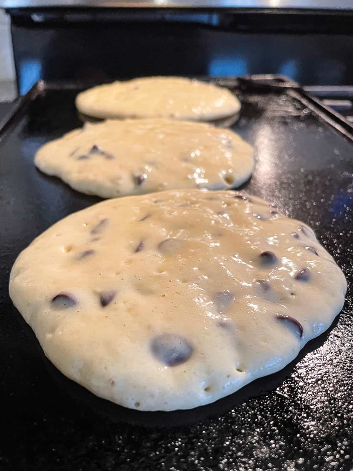 Three gluten-free chocolate chip pancakes cooking on a cast iron griddle. 