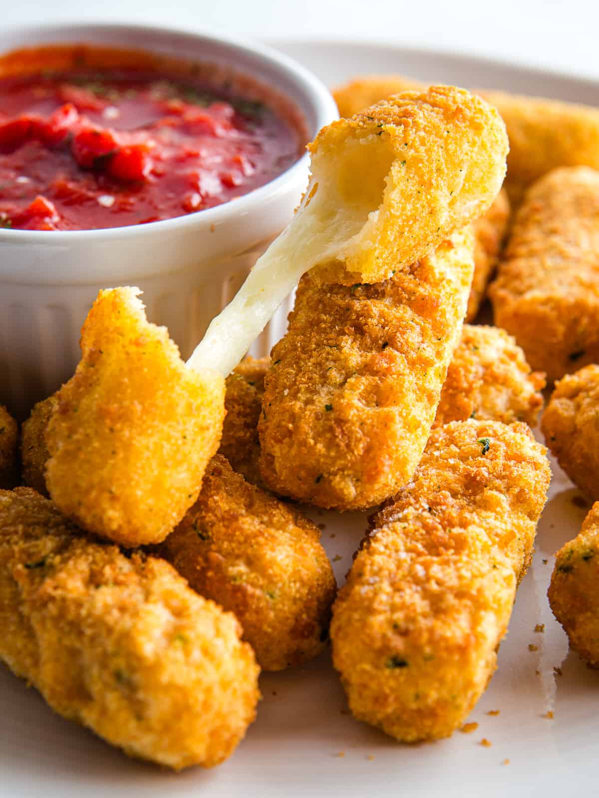 Gluten-free mozzarella sticks on a plate. A small bowl of tomato sauce is in the back.