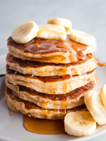 A stack of gluten-free banana pancakes on a plate topped with maple syrup, banana slices, and butter.