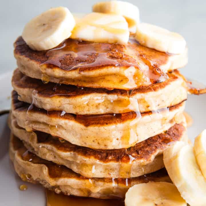A stack of gluten-free banana pancakes on a plate topped with maple syrup, banana slices, and butter.