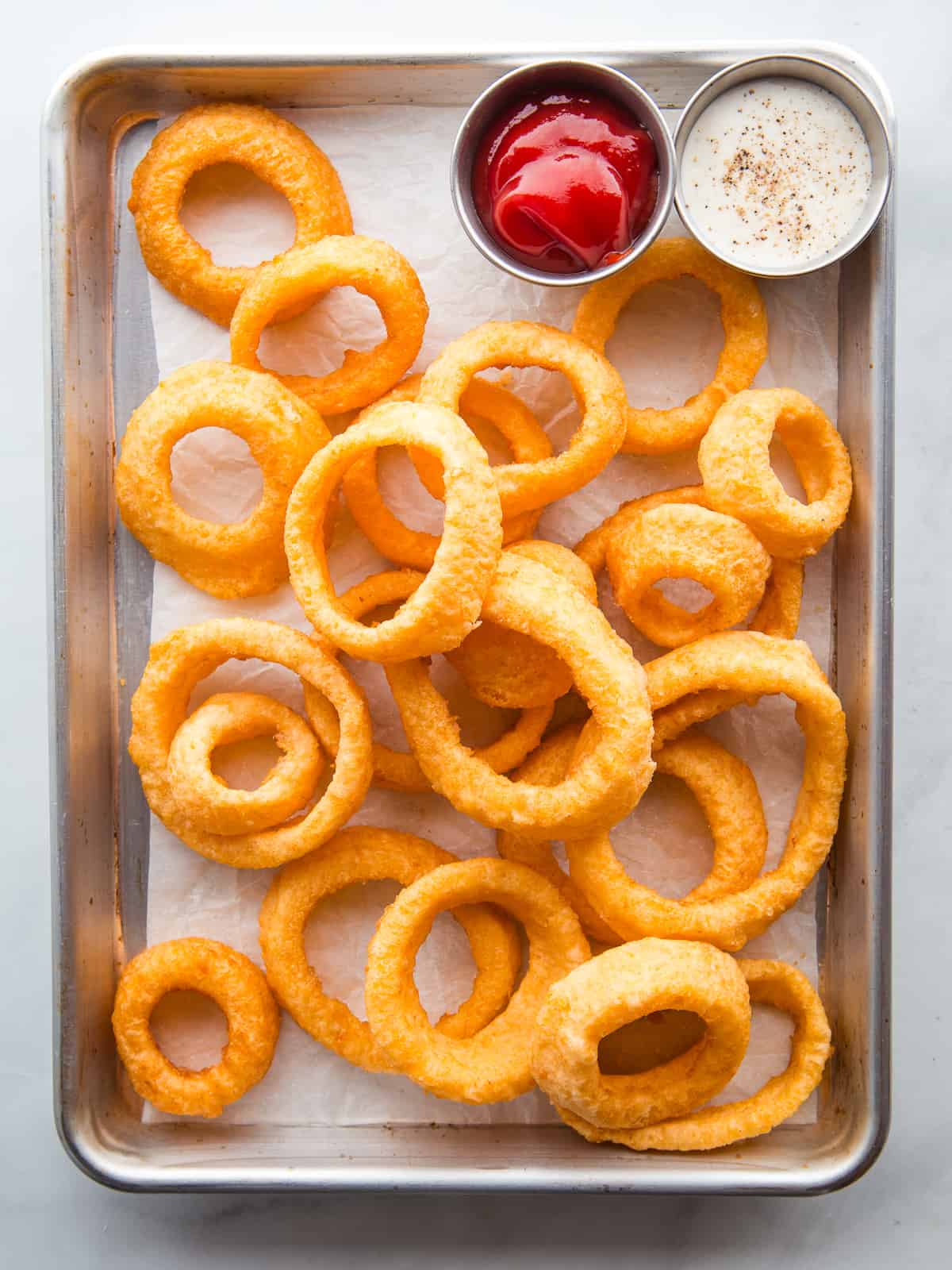 Gluten-free onion rings, along with ketchup and ranch dressing, on a baking sheet.