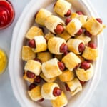 Gluten-free pigs in a blanket on a white platter with ketchup and mustard in small bowls off to the side.