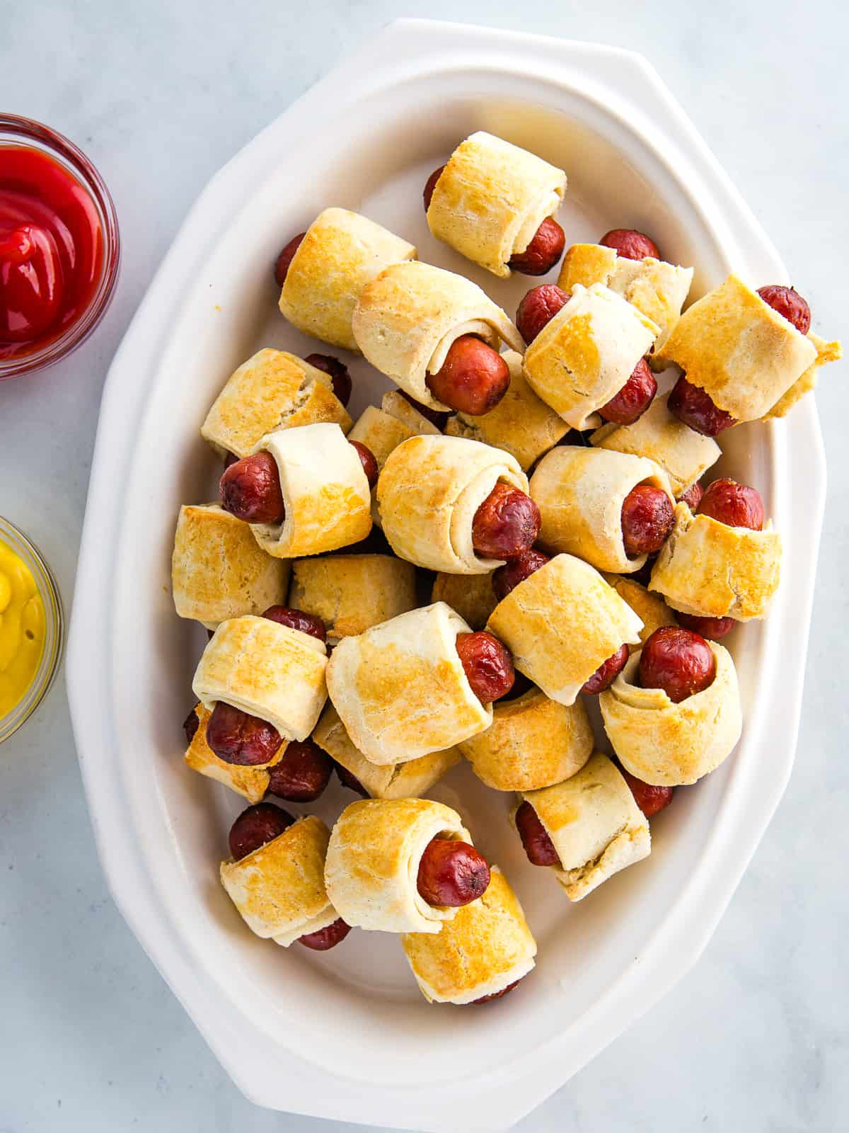 Gluten-free pigs in a blanket on a white platter with ketchup and mustard in small bowls off to the side.