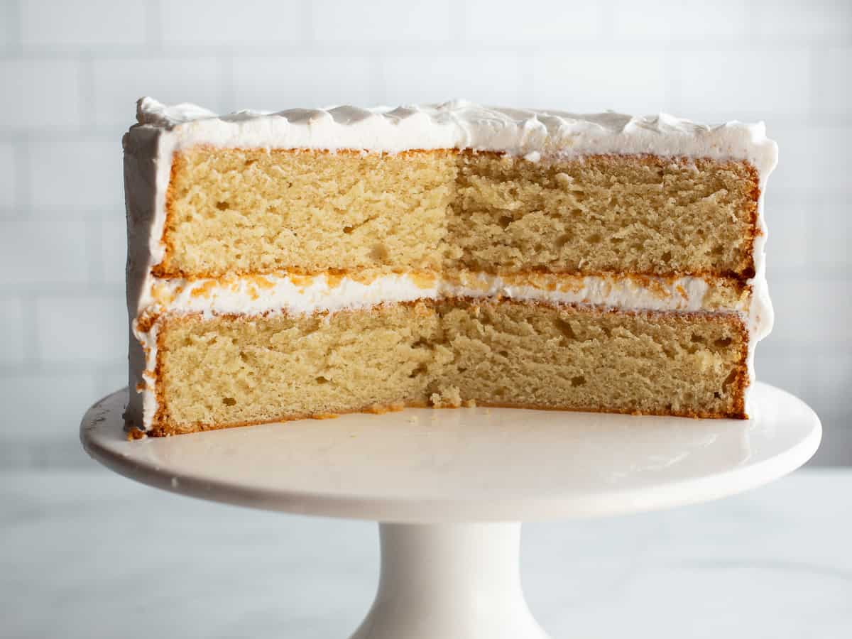 Gluten-free vanilla cake on a white cake stand. The cake is cut, showing the inside crumb and vanilla buttercream filling.