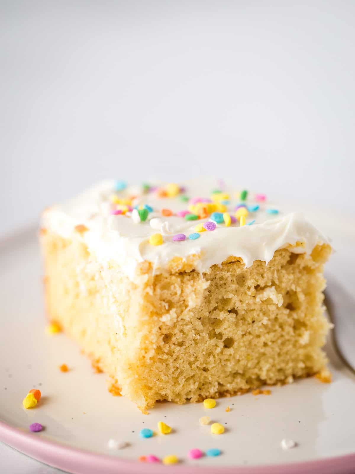 A slice of gluten-free sheet cake on a plate. The cake is frosted with vanilla frosting and sprinkles.