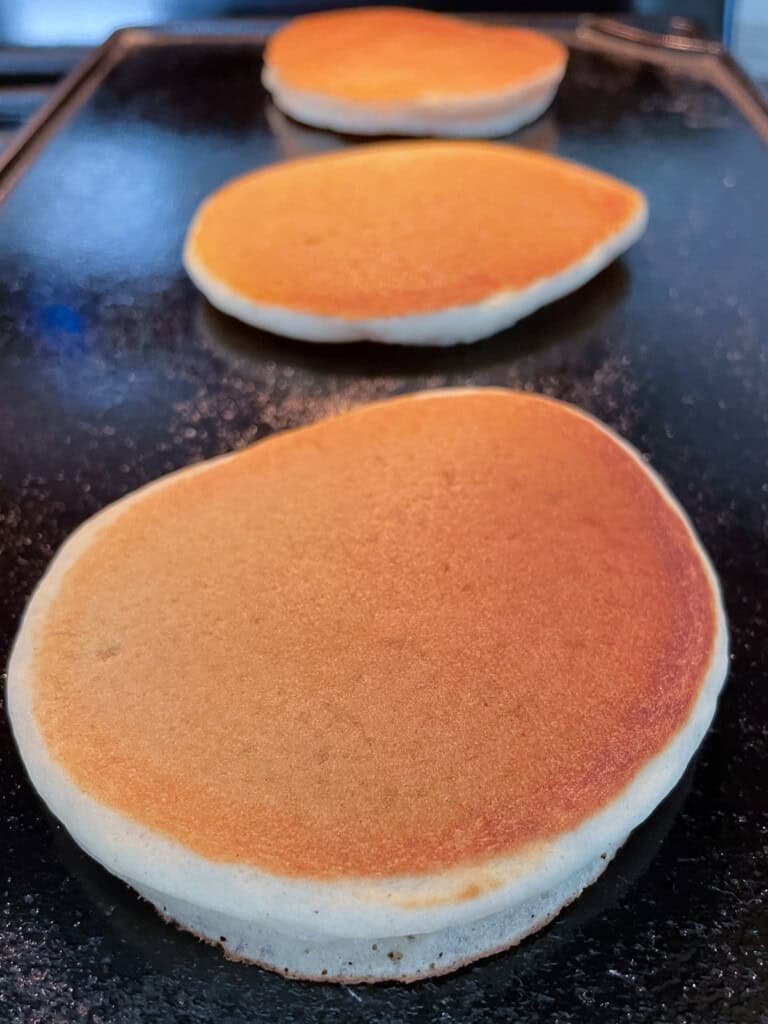 Three gluten-free pancakes cooked on a griddle. The top of the pancakes are brown.