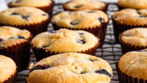 Gluten-free blueberry muffins on a cooling rack. One is unwrapped to show the texture.