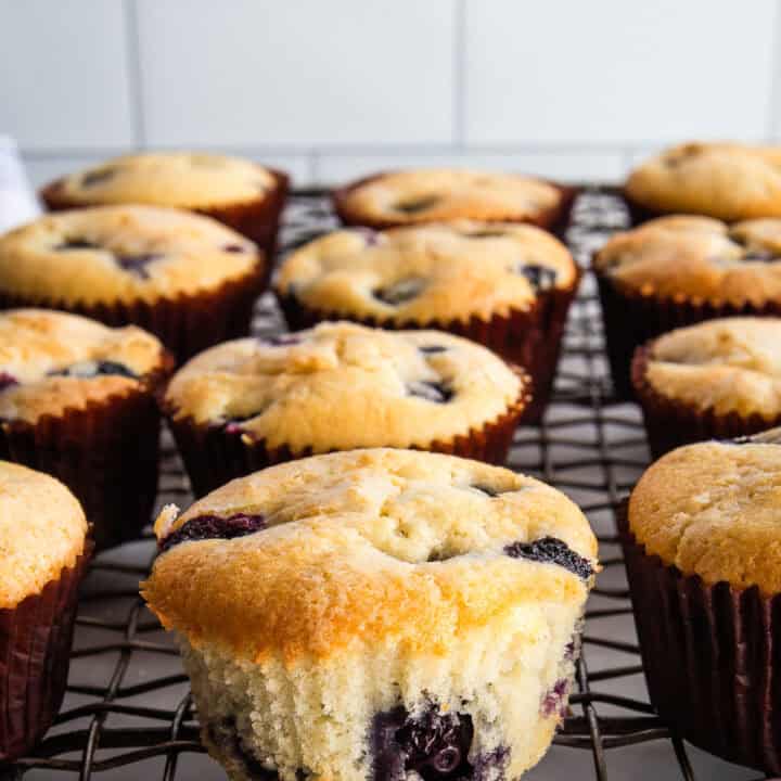 Gluten-free blueberry muffins on a cooling rack. One is unwrapped to show the texture.
