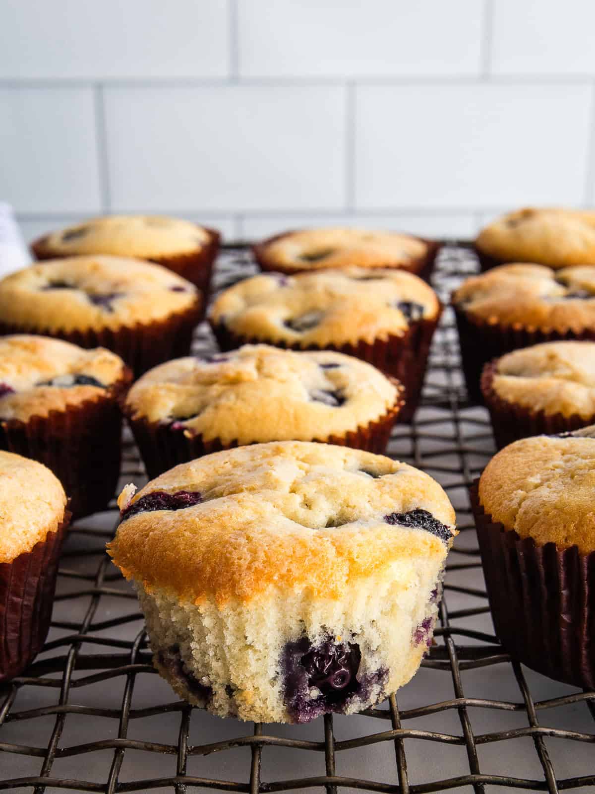 Gluten-free blueberry muffins on a cooling rack. One is unwrapped to show the texture.