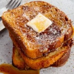 Three slices of French toast on a plate. The toast is topped with butter, syrup, and powdered sugar.