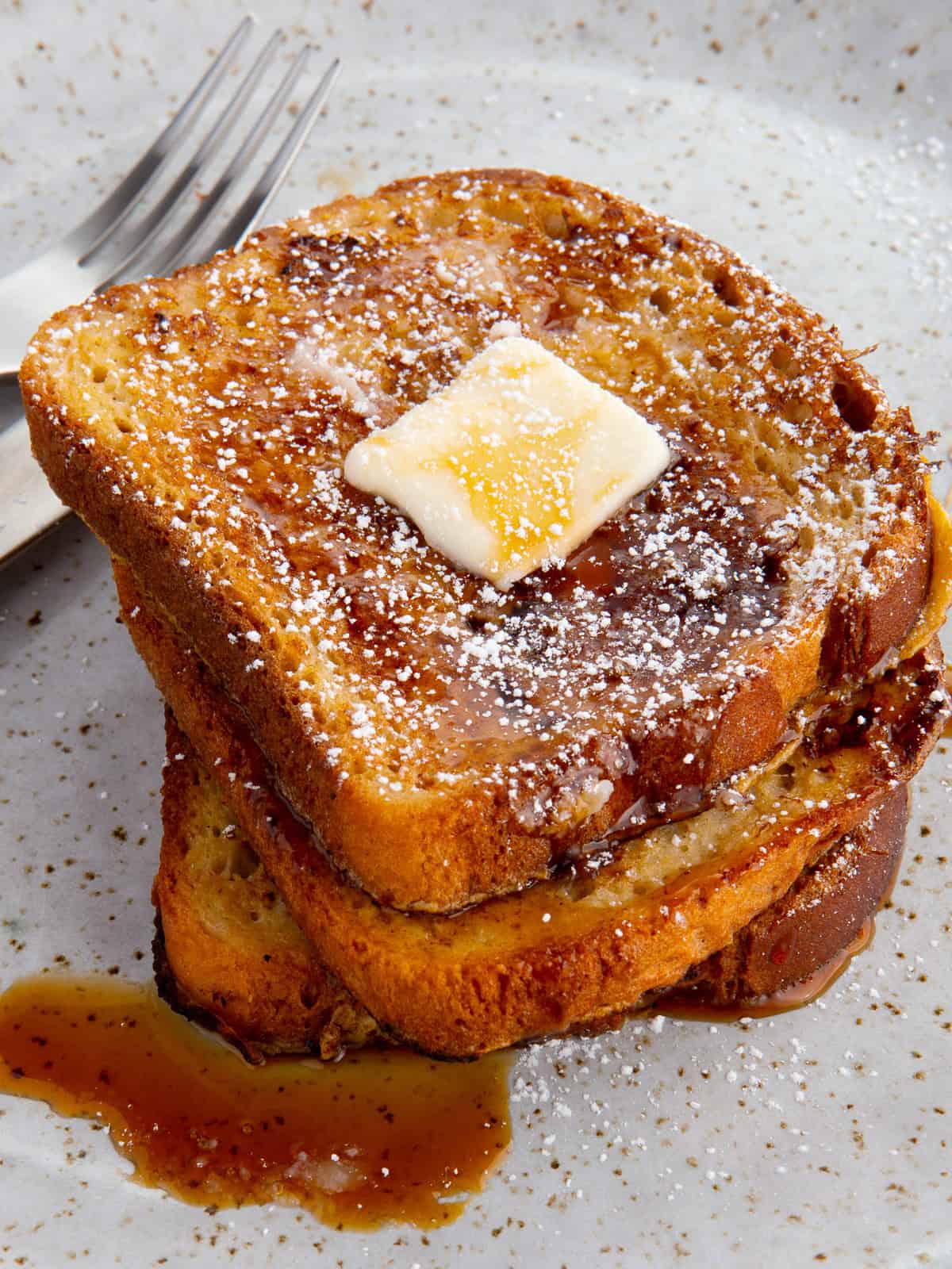 Three slices of French toast on a plate. The toast is topped with butter, syrup, and powdered sugar.