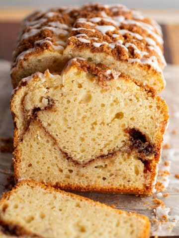Gluten-free cinnamon swirl bread, topped with cinnamon sugar and a vanilla glaze, sits on a cutting board. Two slices are cut from the bread to show the layer of brown sugar and cinnamon in the center.